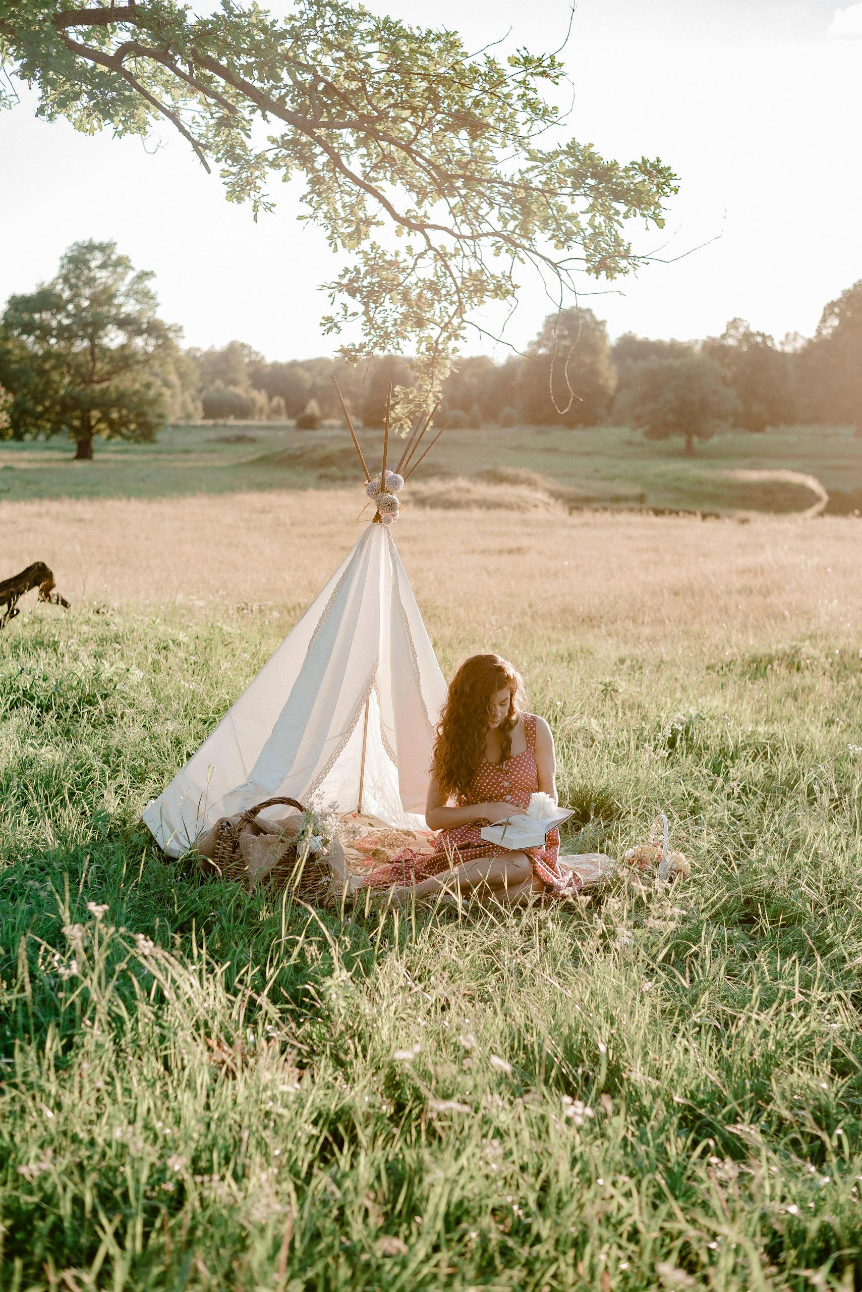 A woman enjoying a peaceful moment reading near a tent in a sunlit grass field.